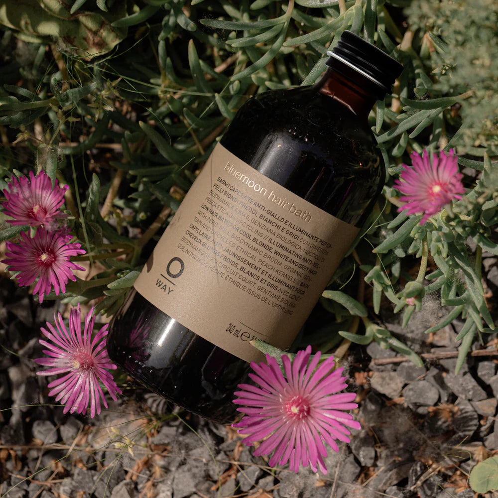Bottle of 'Oway' Blue Moon hair bath on a natural background with pink flowers and green leaves.